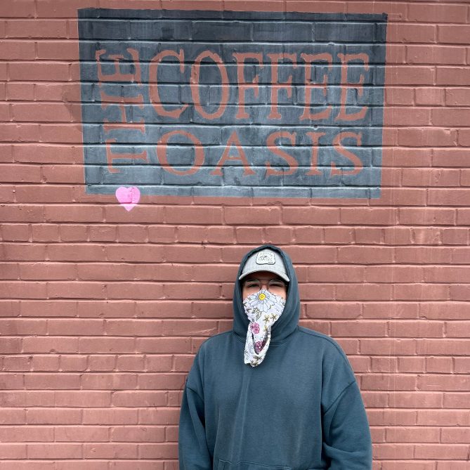 A youth wearing a mask and hooded sweater stands in front of a brick wall at The Coffee Oasis.